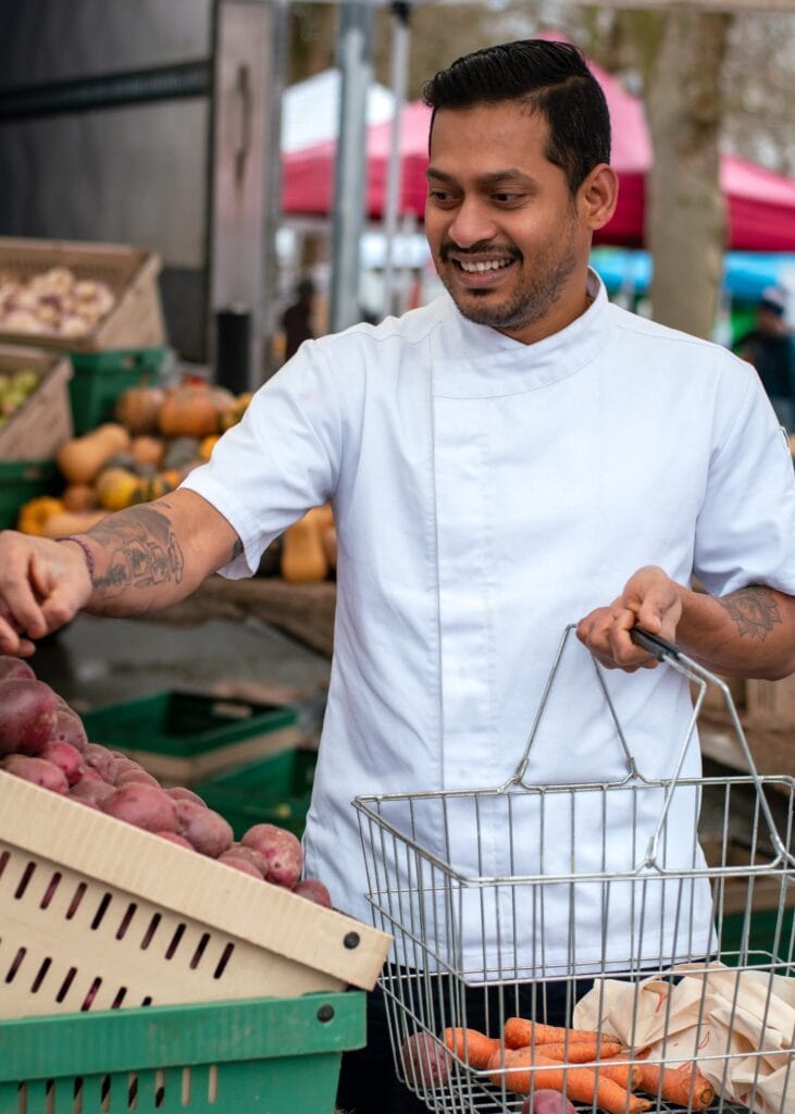 A smiling man in white chef attire shopping for fresh vegetables at an outdoor Indian market, surrounded by pumpkins, potatoes, and carrots, representing authentic Indian ingredients shopping.