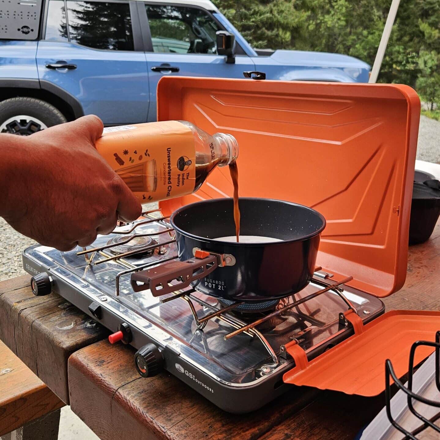 A person pouring chai tea into a pot on a portable stove during an outdoor dining experience.