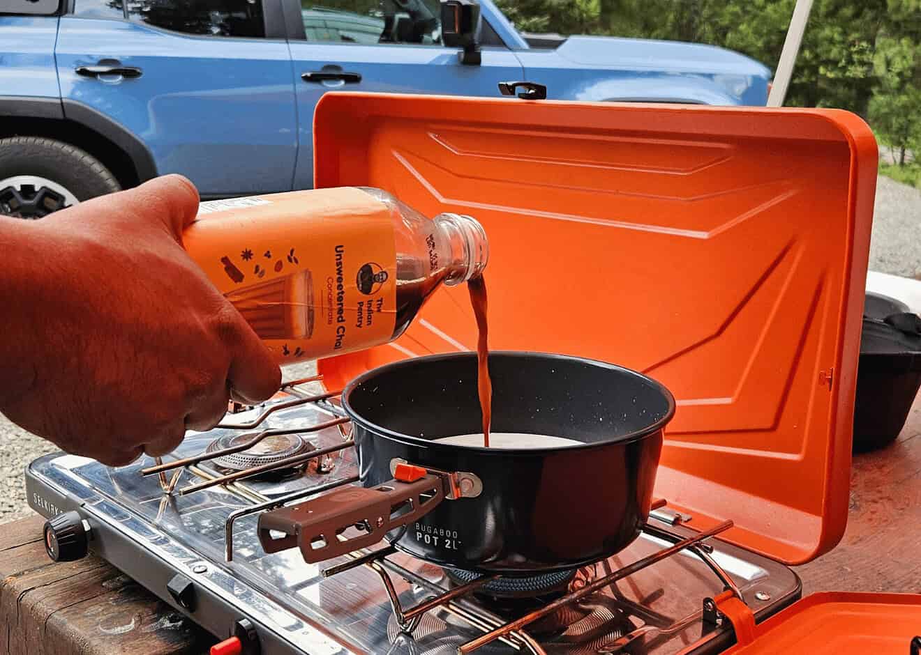 A person pouring traditional Indian chili paste into a black cooking pot on a portable camping stove, outdoors, with a vehicle in the background, showcasing Indian culinary essentials.