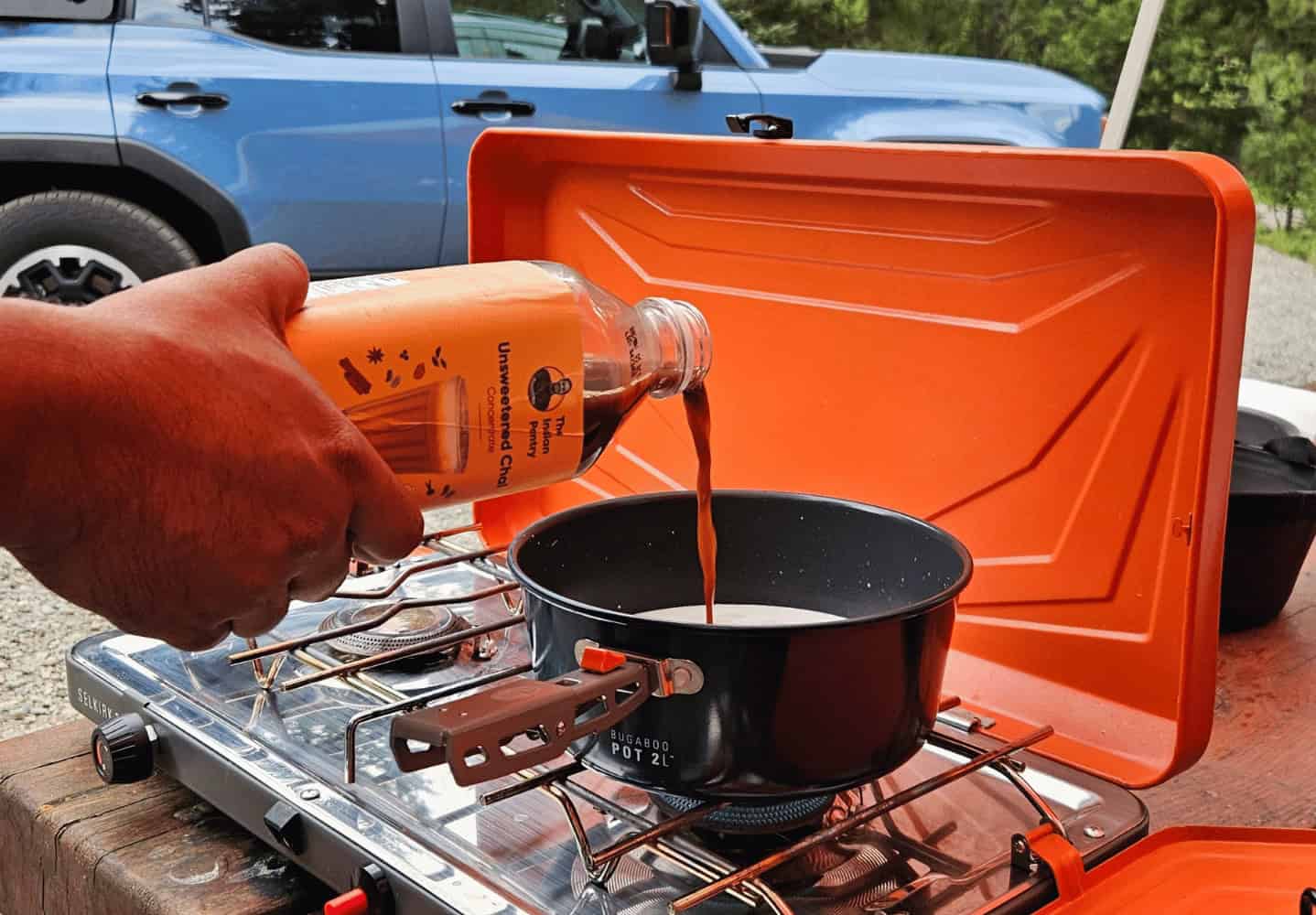 A person pouring traditional Indian chili paste into a black cooking pot on a portable camping stove, outdoors, with a vehicle in the background, showcasing Indian culinary essentials.